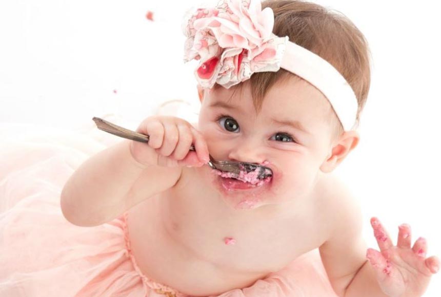 Baby with a spoon in a pink dress and head band at a cake smash