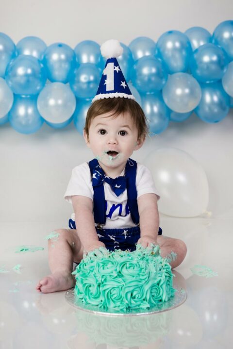 little boy sat with a cake and blue balloons in the background at a cake smash photoshoot