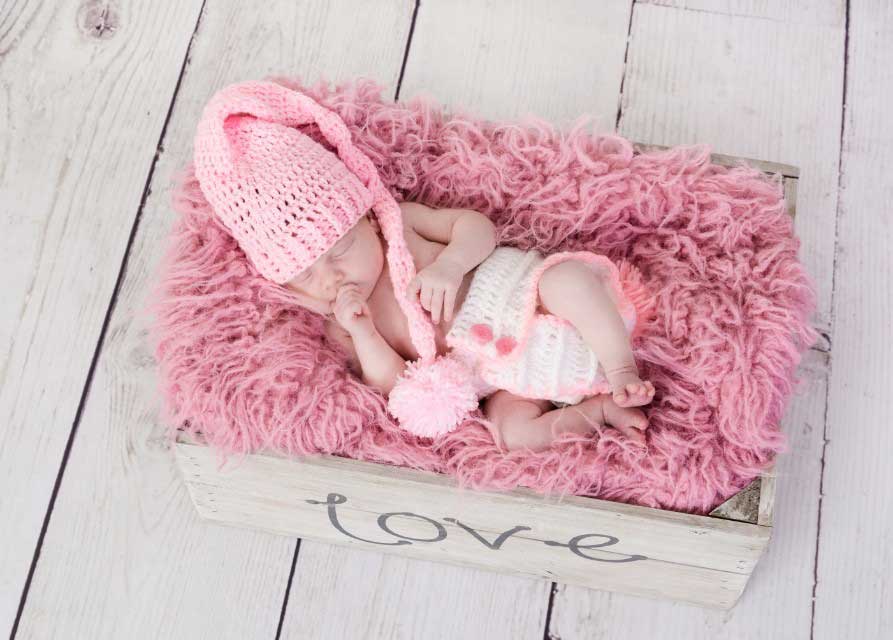 Newborn baby pictured in a crate with the word love on the side with a pink hat and woolly background for a newborn photography photoshoot idea